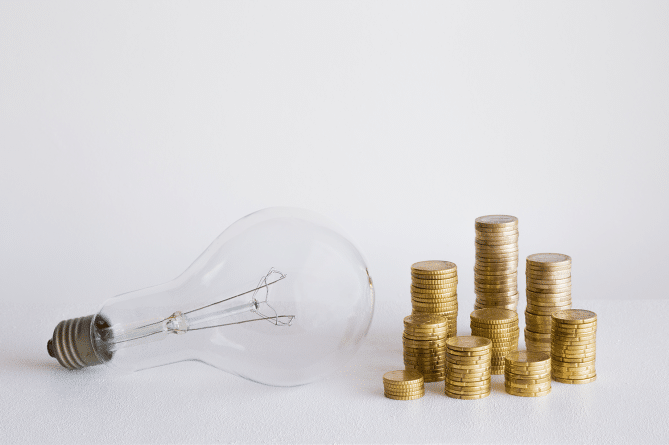 A light bulb next to a pile of gold coins on a white background, signifying a high electric bill.