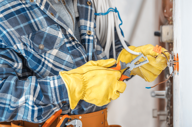 Electrician with yellow gloves and blue flannel performing electrical maintenance for a company.